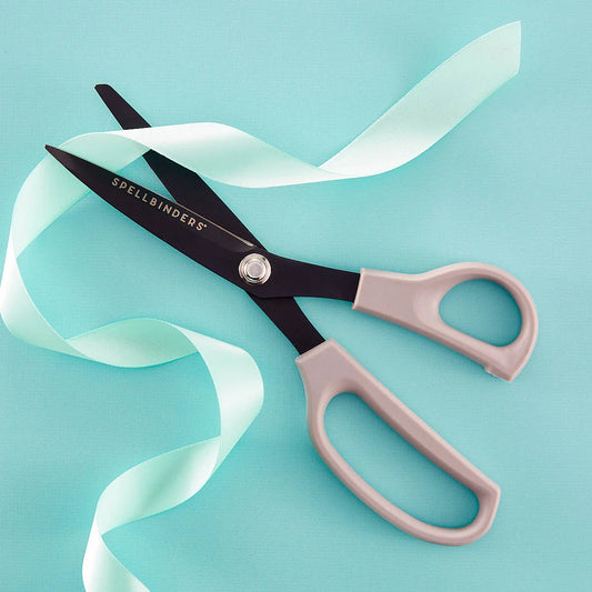 Spellbinders scissors with black blades and pale pink handles, laid diagonally on a light blue background with a pale mint ribbon draped across them.