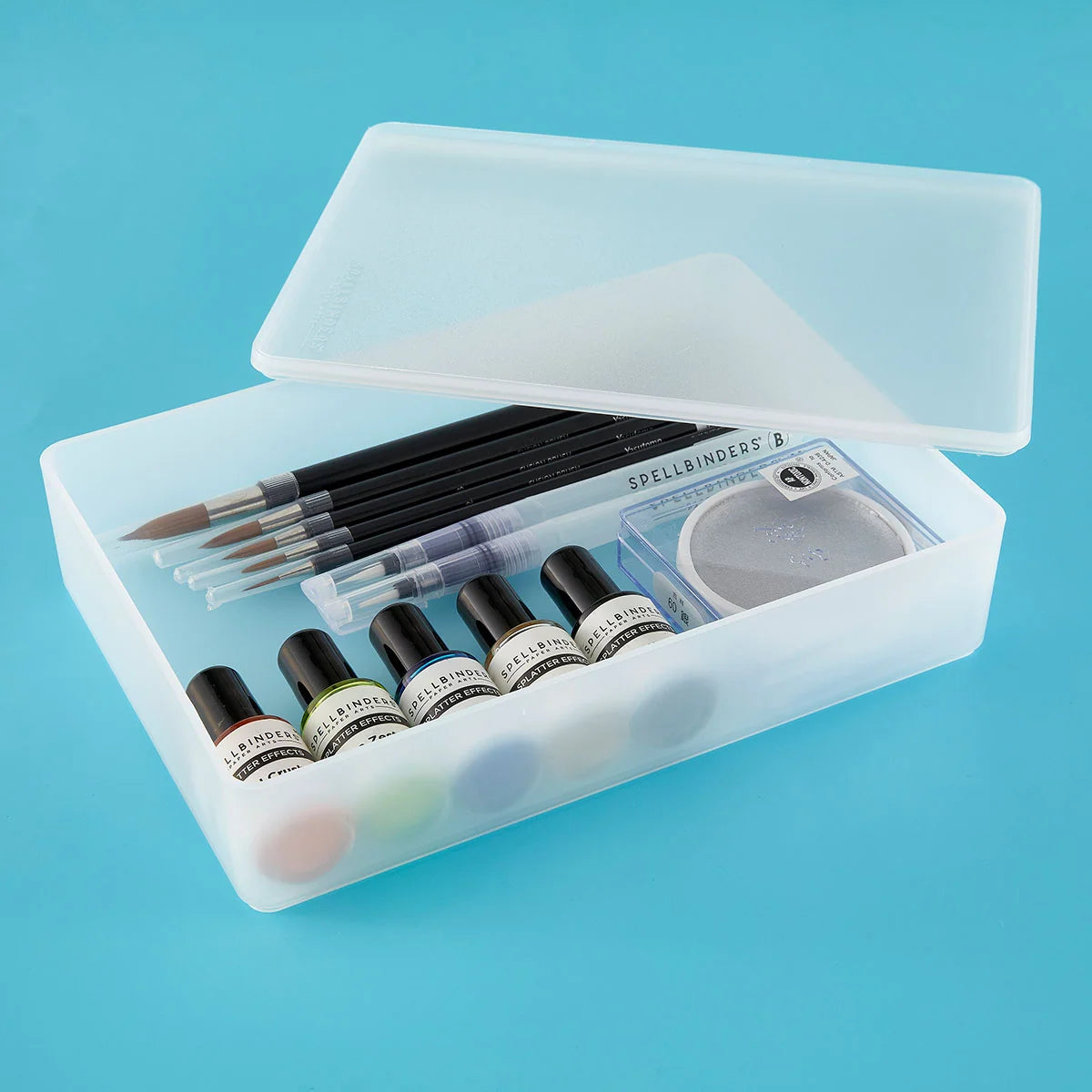 A translucent plastic makeup storage case with its lid open, containing several small cosmetic bottles, a small palette, and two applicator brushes, set against a solid light blue background.