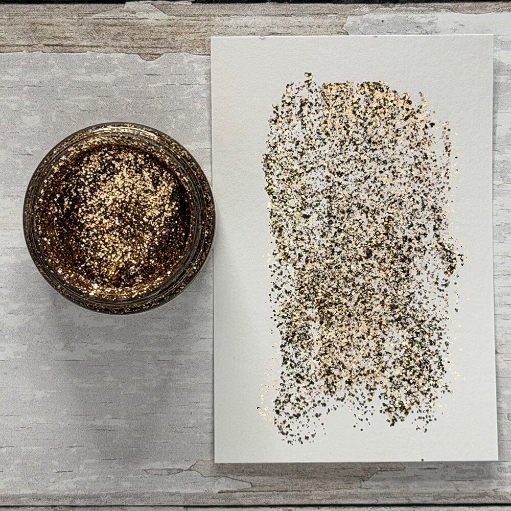 A small round jar filled with coarse gold glitter sits beside a white sheet of paper showing a swatch of the glitter texture, arranged on a light gray wood surface.