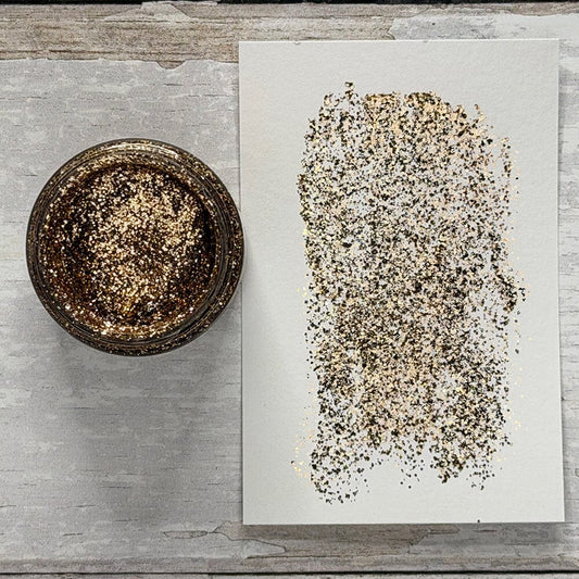 A small round jar filled with coarse gold glitter sits beside a white sheet of paper showing a swatch of the glitter texture, arranged on a light gray wood surface.