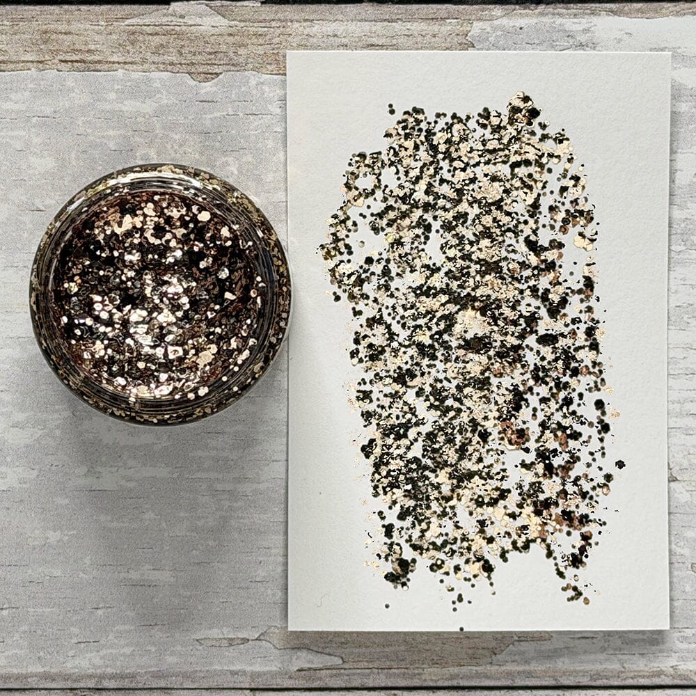 A top-down view of a small round glass jar filled with metallic glitter, placed next to a white sheet of paper showing a swatch of the glitter mix. The background is a rustic, light gray weathered wood surface.
