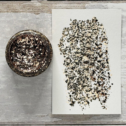 A top-down view of a small round glass jar filled with metallic glitter, placed next to a white sheet of paper showing a swatch of the glitter mix. The background is a rustic, light gray weathered wood surface.
