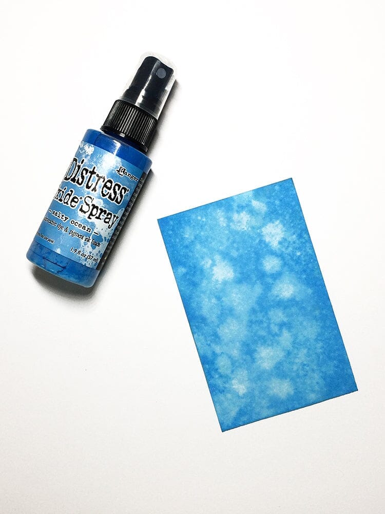 Bottle of Tim Holtz Distress oxide spray next to a blue textured paper on a white background