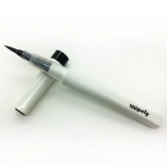 A white and clear brush-tip pen with a black cap, shown with its cap removed and placed behind it on a white background. The handle has the word 'uniquely' printed in black script.