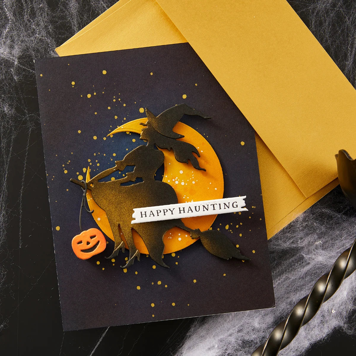 A handmade Halloween greeting card with a black background, featuring a layered design of a witch riding a broomstick in front of a yellow full moon, with gold glitter speckles, a small orange jack-o'-lantern, and a white banner reading 'HAPPY HAUNTING'. A matching yellow envelope is placed behind the card on a dark, cobweb-covered surface.