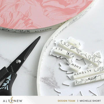 White stamps with floral designs on a white surface with a pair of scissors and a pink container in the background.