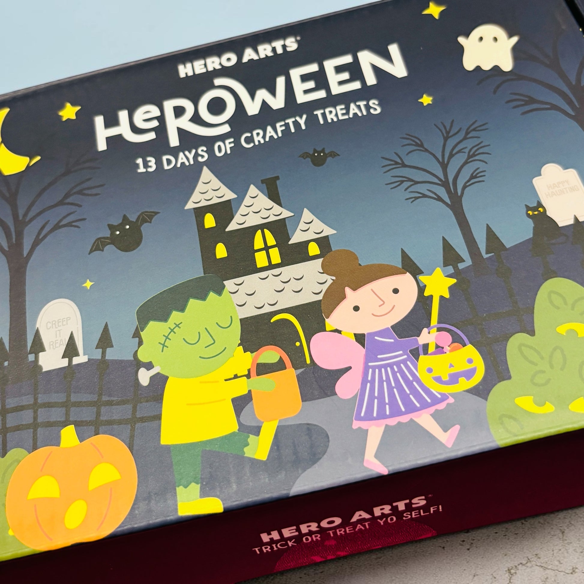 A boxed product labeled HERO ARTS Heroween: 13 Days of Crafty Treats, shown at an angle. The box features Halloween artwork with a haunted house, pumpkins, ghosts, bats, and two children in costume.