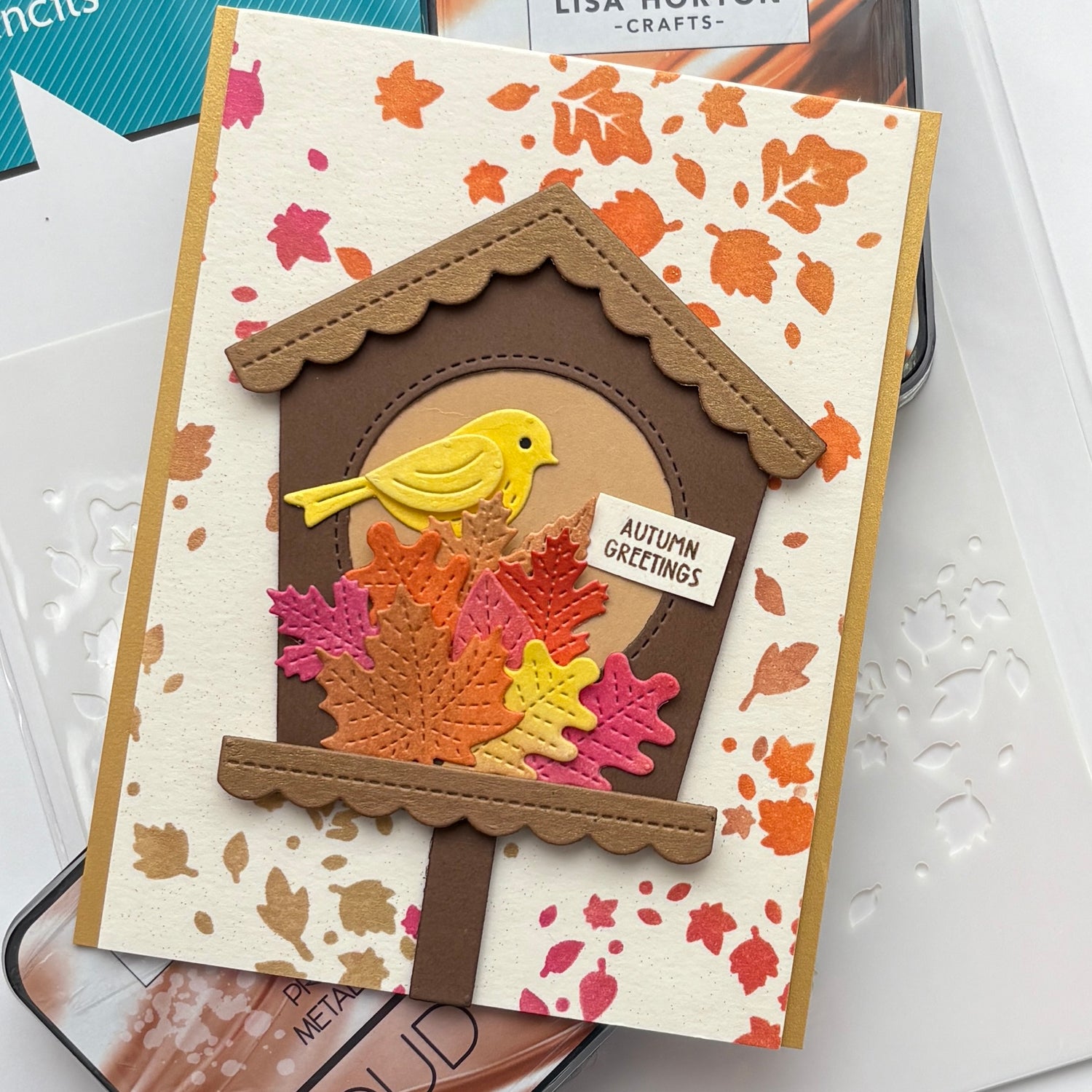 A handmade autumn-themed greeting card featuring a brown birdhouse with a yellow bird, layered with orange, pink, and gold foil leaves on a cream background with scattered leaf prints. A small tag reads 'AUTUMN GREETINGS'.