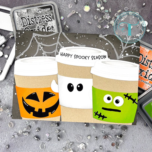 A handmade Halloween-themed coffee cup card featuring three spooky coffee cups with jack-o'-lantern, cute eyes, and Frankenstein's stitched face, displayed on a gray textured background with craft supplies.